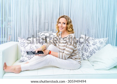 Smiling elegant woman sitting on a sofa in a living room and watching TV. Home interior, furniture. Lifestyle.