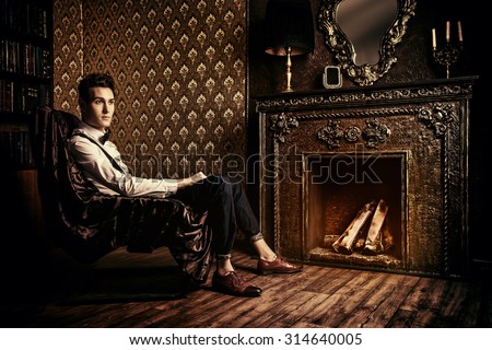 Elegant handsome young man sitting by the fireplace in a room with classic vintage interior. Fashion shot.