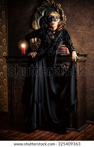 Charming mysterious girl in black mask and black medieval dress stands in a castle living room. Vampire. Halloween concept. Vintage style.