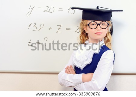 Cute smart schoolgirl in big glasses and academic hat stands at the blackboard. Education.
