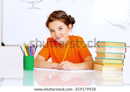 Pretty smart schoolgirl sitting at the desk in the classroom on blackboard background. Education.