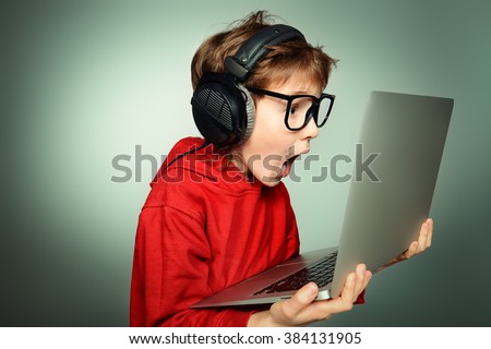 Cute boy in spectacles and headphones looking at his laptop monitor and shouting. Studio shot.