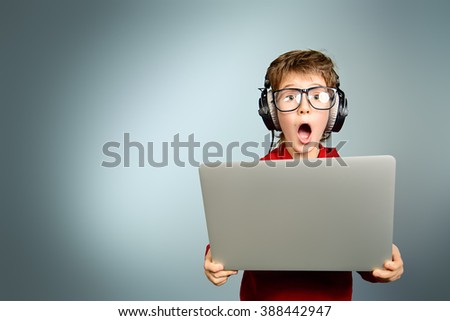 Cute boy in spectacles and headphones looking at his laptop monitor and shouting. Studio shot.