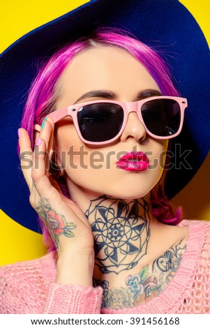 Pretty girl with crimson hair wearing bright clothes and sunglasses posing over yellow background. Bright style, fashion. Optics style. Tattoo.