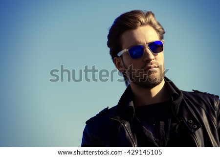 Confident handsome man in sunglasses and leather jacket over blue sky. Men\'s beauty, fashion. Outdoor portrait.