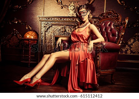 Charming elegant woman in beautiful red dress and masquerade mask is sitting in a chair in a luxury apartment. Classic vintage interior. Beauty, fashion. Retro style.