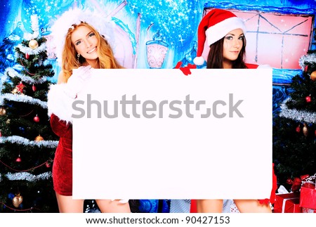 Two sexy young women in Christmas clothes holding white board over Christmas background.