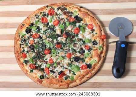 Mediterranean style pizza with chicken and various vegetables on a wooden cutting board with cutter on the side