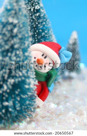 winter scene in glitter, a snowman  is playing hide and go seek, peeking or hiding  behind miniature pine trees