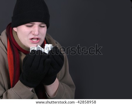Teenage male sneezing into a tissue dealing with the winter flu, dressed in outerwear like gloves, scarf, and wool hat on a grey background