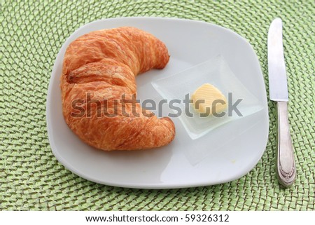Golden croissant with orange slice on a white plate with a knife and a pat of butter on a green wicker placemat