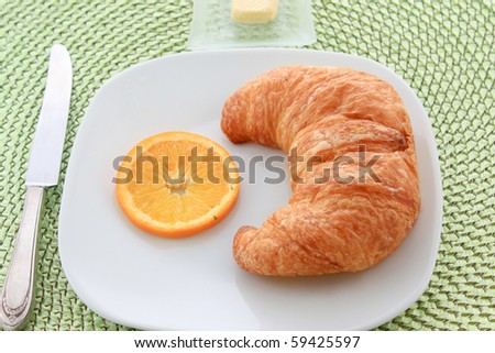 Golden croissant with orange slice on a white plate with a knife and a pat of butter on a green wicker place mat