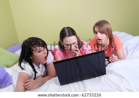 Group of three teenage girls with pigtails and braided hair having a slumber party or sleepover laying on a bed reacting to screen on laptop