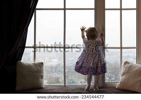 A girl standing near the window, watching  outside, where is the rainy day