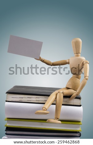 Wooden mannequin sitting on stack of books holding white business card.