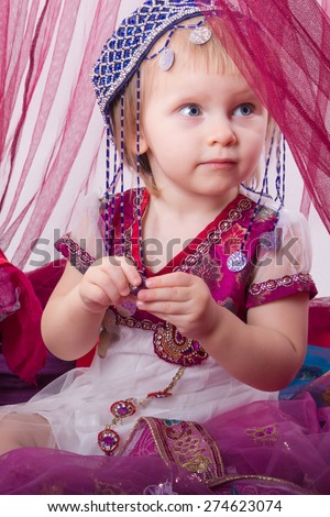 Little young east kid child  girl, sultana, Princess in Indian dress sari burqa scarf veil holding a pearl necklace sitting on a floor pillow at home interior under pink canopy. Childhood, copy space