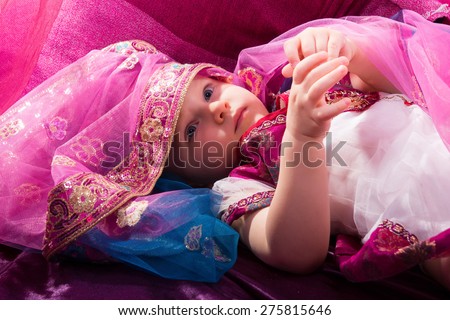 Little young east girl, sultana, Princess in Indian dress sari burqa scarf veil holding a pearl necklace sitting on a floor pillow at home interior under pink canopy. Childhood, copy space, doll house
