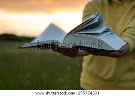Female hands holding opened hardback book, diary with fanned pages on blurred nature landscape background against sunset sky with back light. Copy space, back to school education concept.
