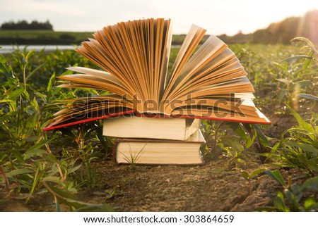 Opened hardback book diary, fanned pages on blurred nature landscape backdrop, lying in summer field on green grass. Books stacking. Copy space, back to school education background.