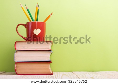 Composition with old vintage hardback books and red cup with yellow heart shape and pencils on wooden deck table and green background. Books stacking. Back to school. Copy Space. Education background.