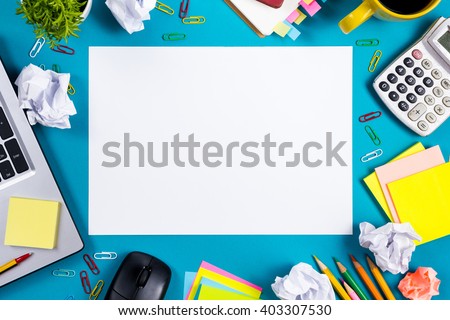 Office table desk with set of colorful supplies, white blank note pad, cup, pen, pc, crumpled paper, flower on blue background. Top view and copy space for text.