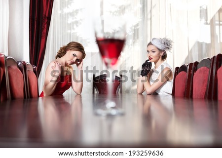 Two women sitting at table in restaurant. A glass of of red wine costs in the center of the table.
