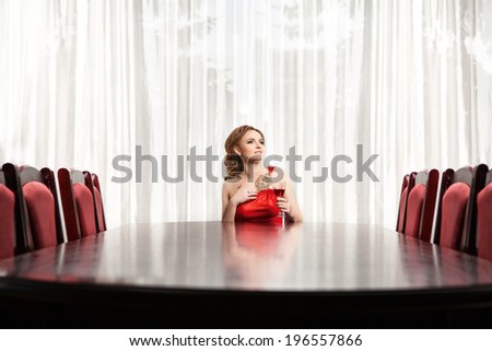 Portrait of a beautiful woman in red dress. One women sitting at table in restaurant. A glass of of red wine costs in the center of the table.
