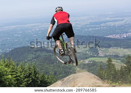 Rear view of mountain bike rider who jumps over a dirt track kicker. The chosen perspective gives the impression of a jump into the precipice. The background shows the black forest in germany.