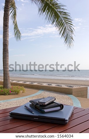 A closed laptop and a personal planner on a table by a beach, exotic mobile office.  Vertical image.