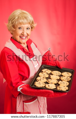 Sweet homemaker grandma holding a tray of fresh baked chocolate chip cookies.  Photographed in front of red background.