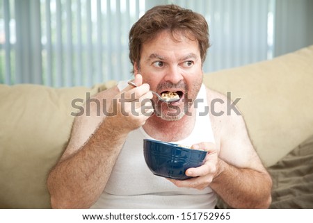 Unemployed man sitting on the couch eating cereal as he watches television.