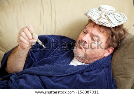 Man in his bathrobe on the couch, home sick from work.  He has an icepack on his head and is taking his temperature.
