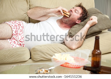 Unemployed middle aged man at home on the couch in his underwear, eating a hamburger,  with a marijuana joint in the ashtray and beer bottles lying around.