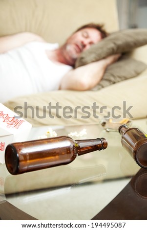 Man asleep on the couch with ashtray, takeout food container, and beer bottles on coffee table.  Shallow Depth of field with focus on beer bottles.