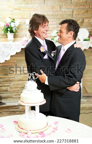 Two handsome gay men in tuxedos embrace with love at their wedding reception.  Wedding cake and rose petals in foreground.