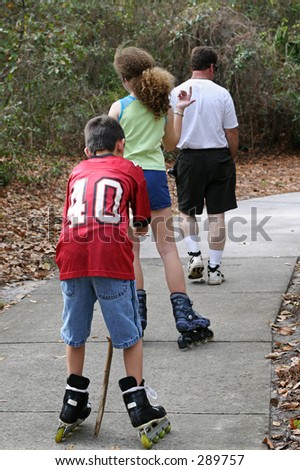 Two teens skating up a path in a park, with an adult walking ahead.