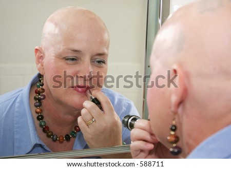 A woman bald from a health problem putting on makeup in the mirror.