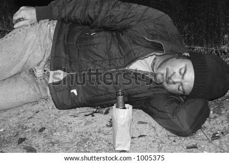 A black and white photo of a homeless man sleeping on the ground by a dumpster.  He has a bottle of wine in a bag beside him.