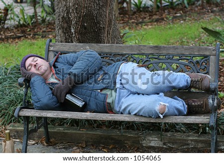 A full view of a homeless man asleep on a park bench with his wine bottle.