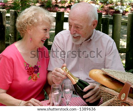 An attractive senior couple on a picnic, looking lovingly into each-others eyes as he opens the wine.