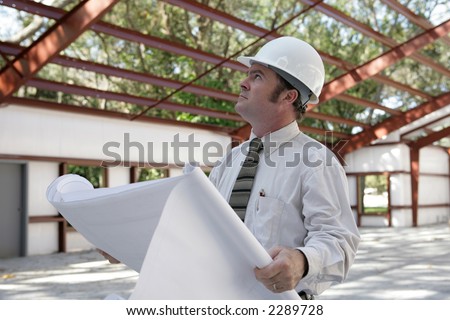 A construction inspector holding blueprints and looking at the roof beams of a steel building in progress.