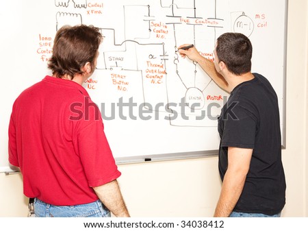 Electrical engineering student draws a diagram of a circuit on the white board while teacher looks on.