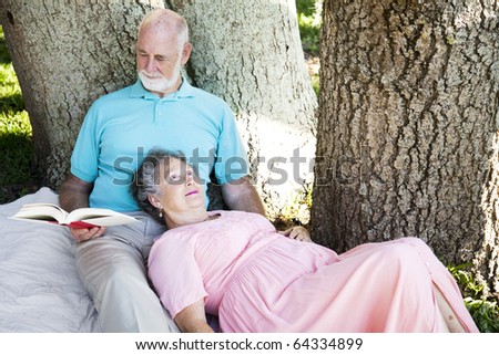 Senior man reads to his wife, as they relax in the park.