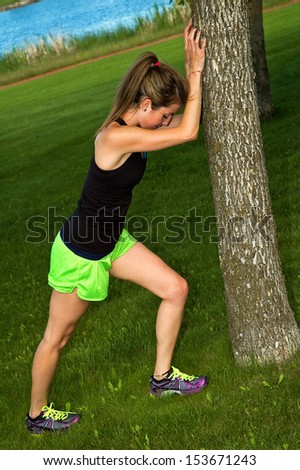 Young woman stretching her calf muscle, against a tree in a city park.