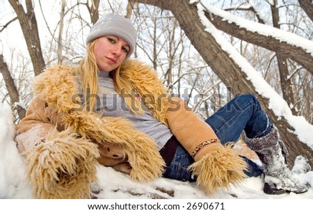 A young woman laying down, wearing a furry tan colored coat in the woods.