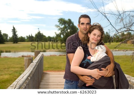 Young family with a new baby, standing on a bridge in a park.