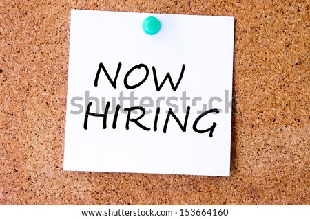 Now Hiring written on an yellow sticky note pinned with blue push pin on cork bulletin board.