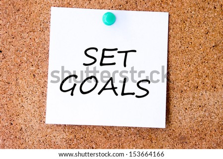 Set Goals on white sticky note pinned with blue push pin on cork board