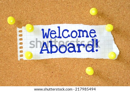 The phrase Welcome Aboard! typed onto a scrap of lined paper and pinned to a cork notice board. A phrase used to welcome a new employee or team member.