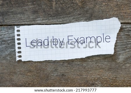 The phrase Lead By Example typed on a paper note on wooden board